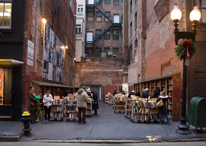 Libreria accogliente all'aperto in un vicolo urbano con persone che sfogliano libri circondate da muri di mattoni e lampioni d'epoca.