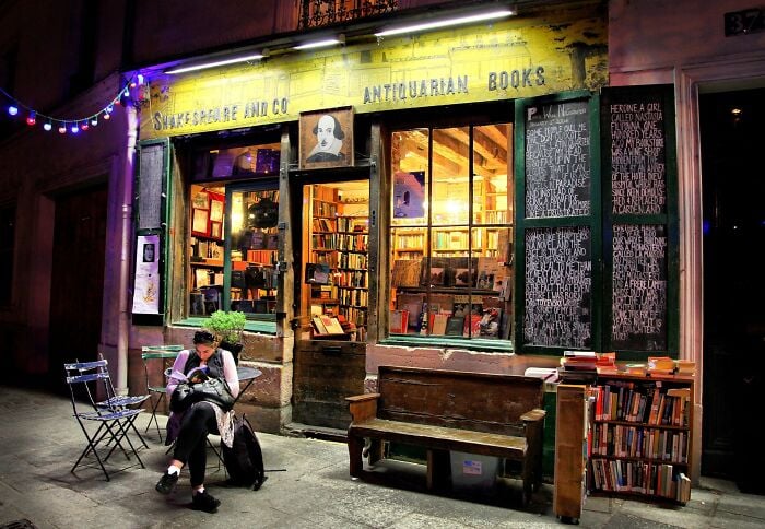 Libreria accogliente e grandiosa di notte con illuminazione calda, piena di libri e una persona che legge fuori.