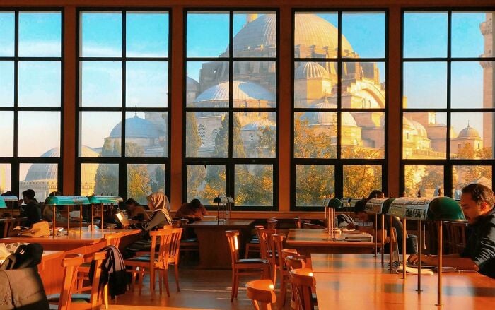 Cozy library interior with large windows showing grandiose architecture, and people reading and studying at wooden tables.