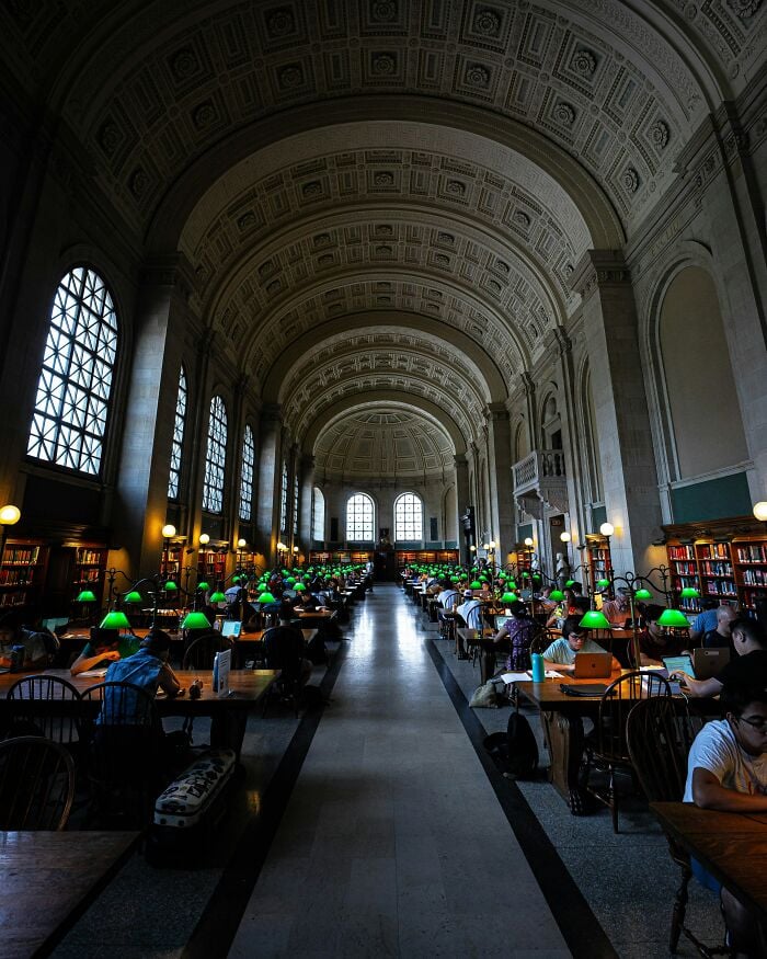 Interni grandiosi della biblioteca con alti soffitti a volta, grandi finestre e file di tavoli da studio con lampade verdi e lettori.