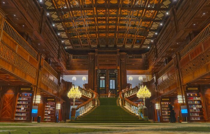 Interior grandioso de biblioteca com uma grande escadaria, paredes de madeira ornamentadas e estantes cheias de livros em um cenário mágico.
